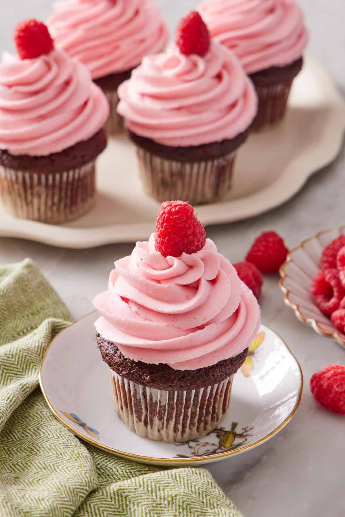 A plate with a cupcake topped with raspberry buttercream frosting and a fresh raspberry. More frosted cupcakes in the background. Fresh raspberries scattered on the surface.