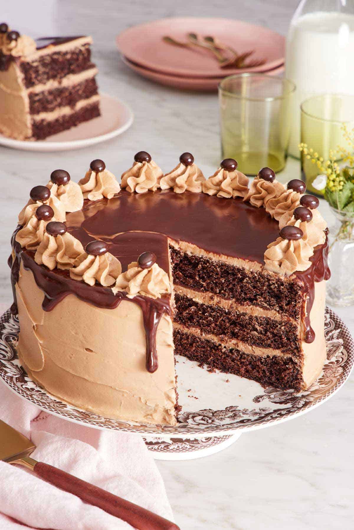 A mocha cake on a cake stand with a slice cut out. A plated slice in the background along with glasses, milk, and plates.
