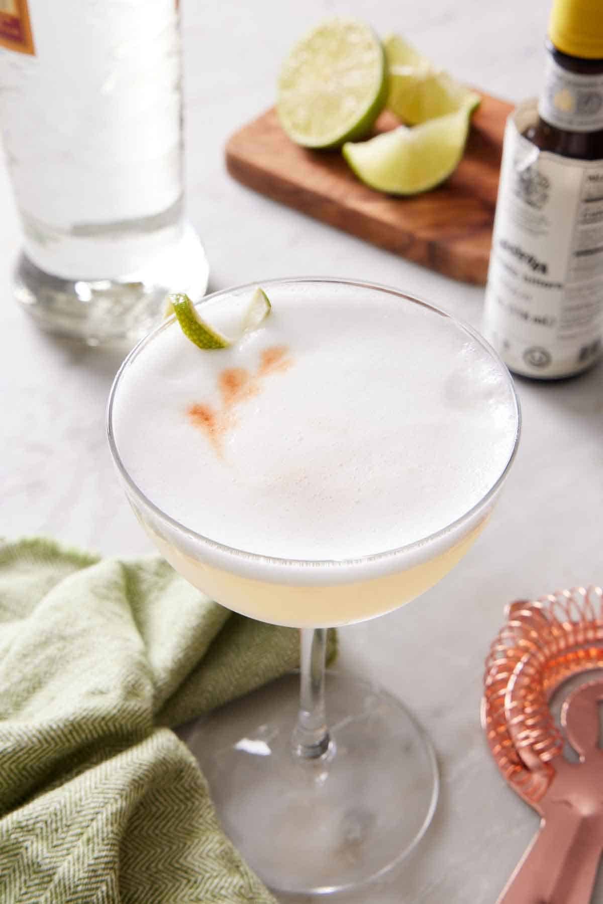 A glass of pisco sour with a lime twist. A bottle of pisco and bitters in the background with cut limes.