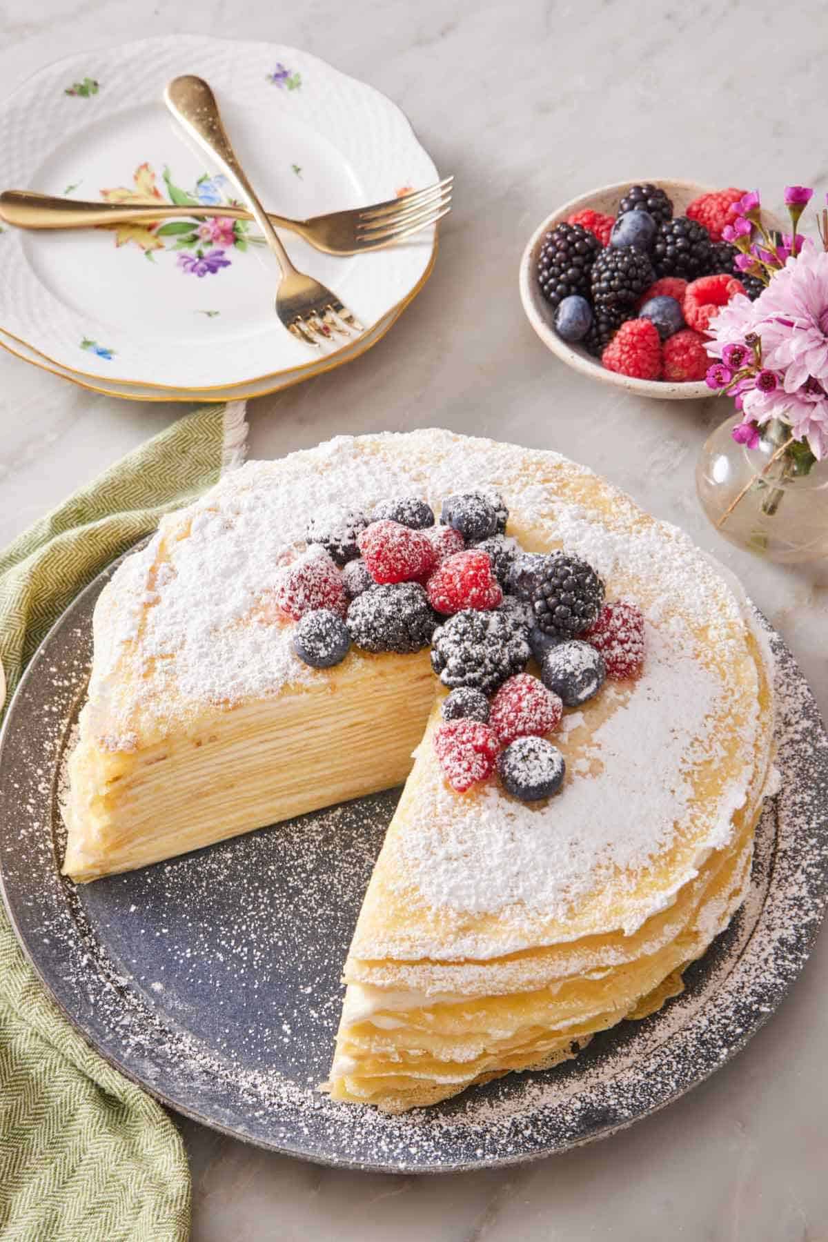 Overhead view of homemade crepe cake with one slice cut out. It is decorated with powdered sugar and mixed fresh berries. A bowl of berries, flowers, and serving plates with forks are on the side.