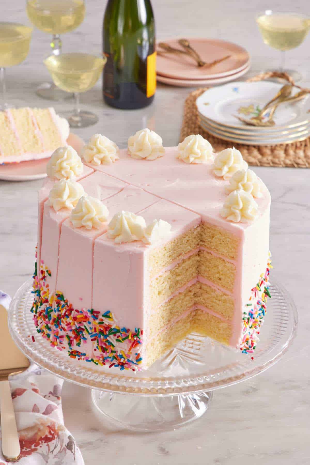 Cake on a cake stand with cuts in it demonstrating how to cut a round cake neatly and easily. Drinks in glasses and serving plates in the background.
