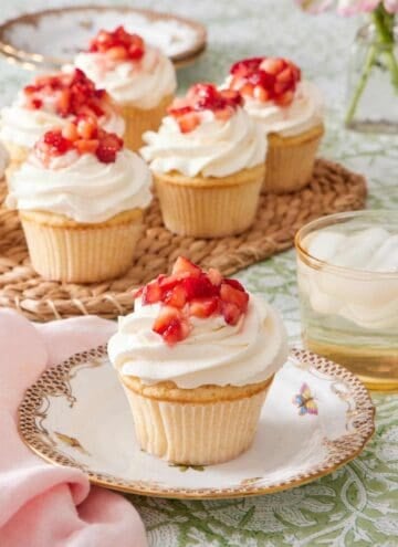 Cupcake on a plate topped with whipped topping and diced strawberries. A drink sits alongside it, and there's more strawberry shortcake cupcakes in the background.