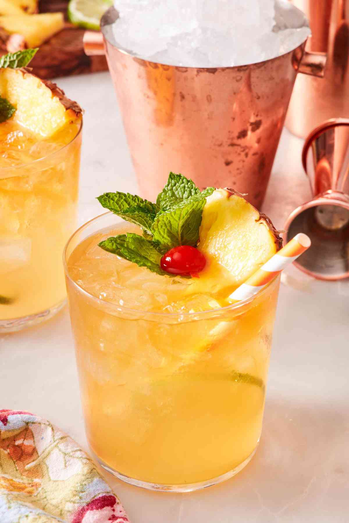 Mai tai garnished with pineapple, mint, and cherry, with a cute straw. A cocktail shaker full of ice in the background.