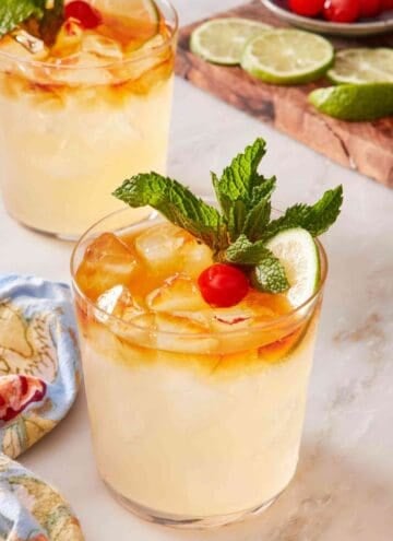 Easy mai tai recipe garnished with lime, mint, and cherry. Another cocktail and a wooden board with cocktail garnishes in the background.
