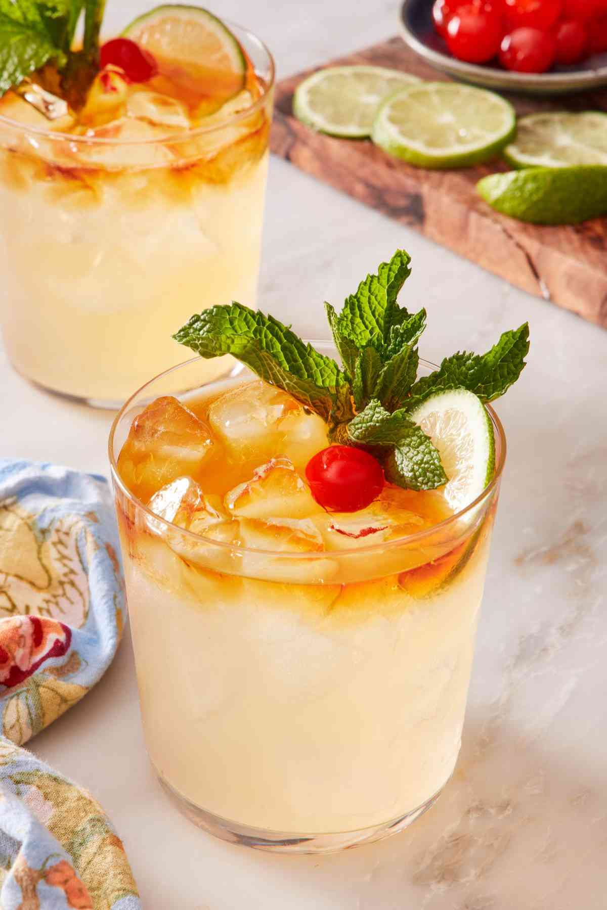 Easy mai tai recipe garnished with lime, mint, and cherry. Another cocktail and a wooden board with cocktail garnishes in the background.