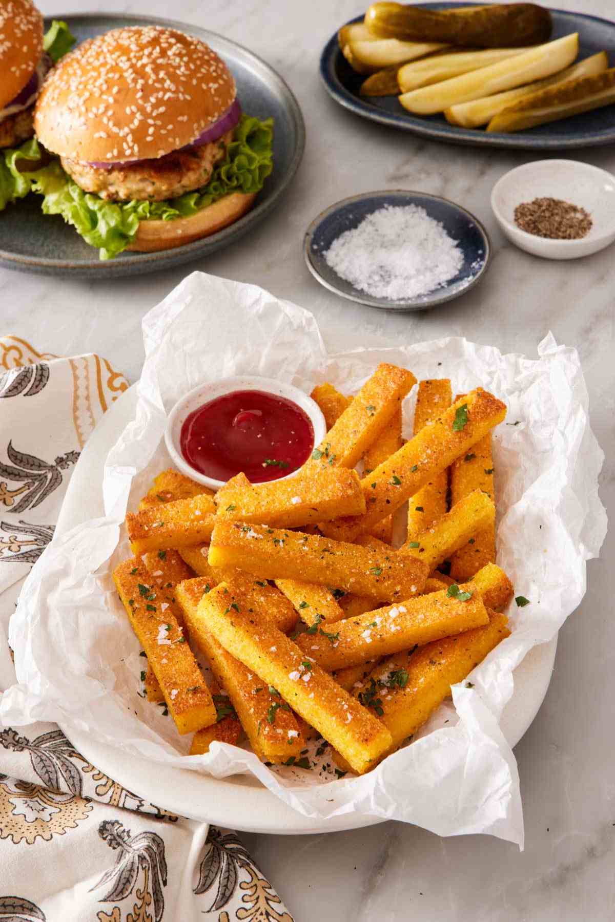 A dish of polenta fries on a paper towel with a burger in the background.