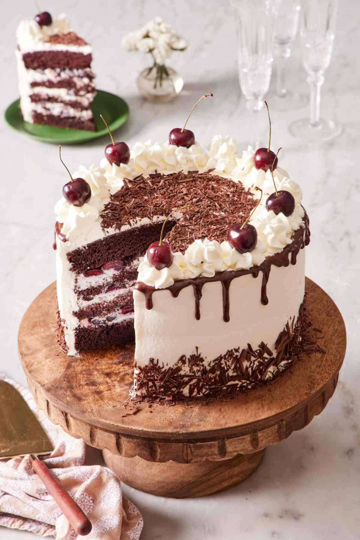 A black forest cake on a wooden cake stand with a slice cue out. The slice is on a small serving plate in the background.