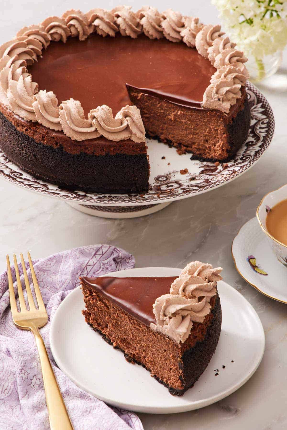 A slice of chocolate cheesecake on a small plate with the rest of the cheesecake on a cake stand right behind it.