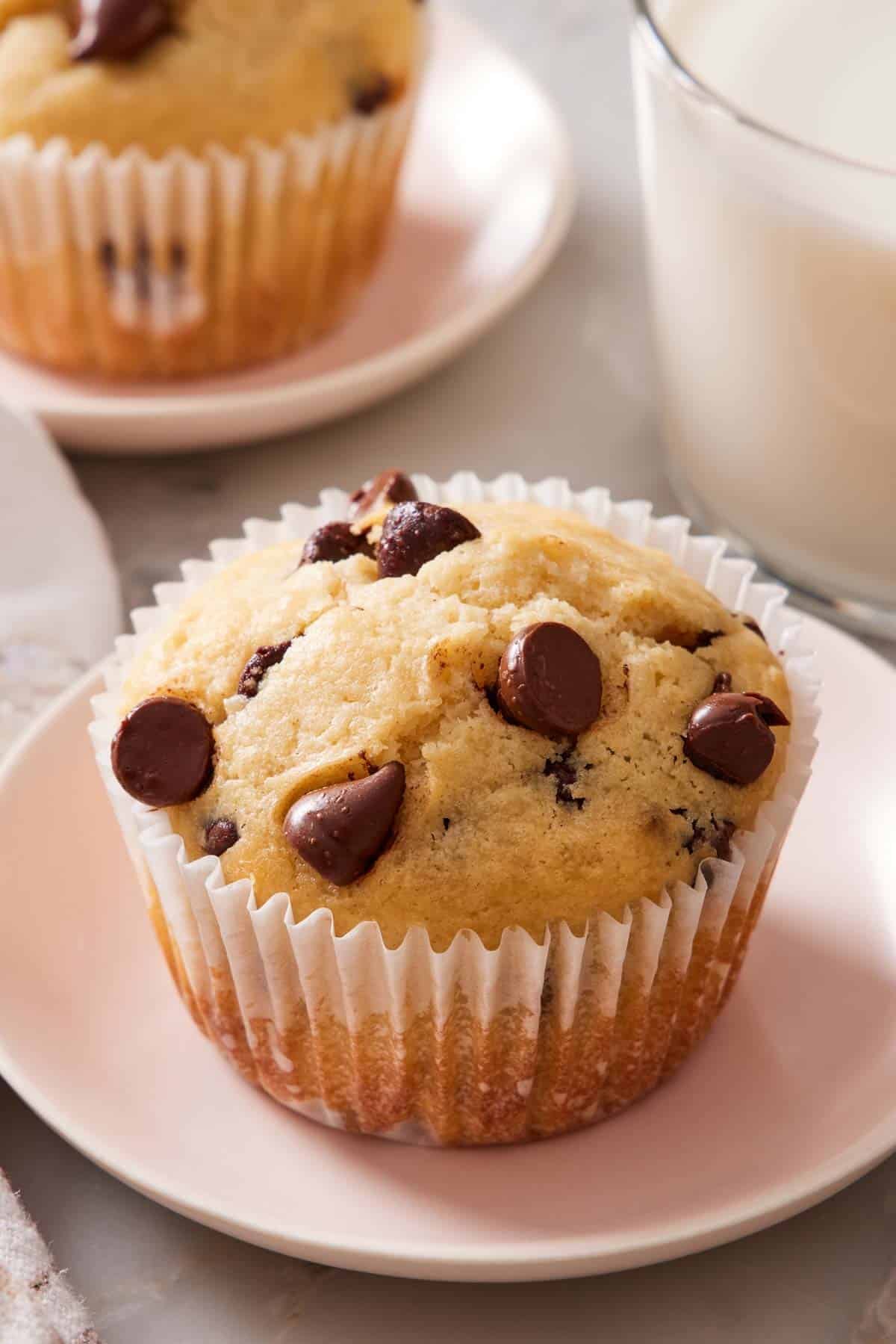 Close-up of a choc chip muffin on a small plate with a glass of milk and another muffin out of focus and mostly out of frame behind it.