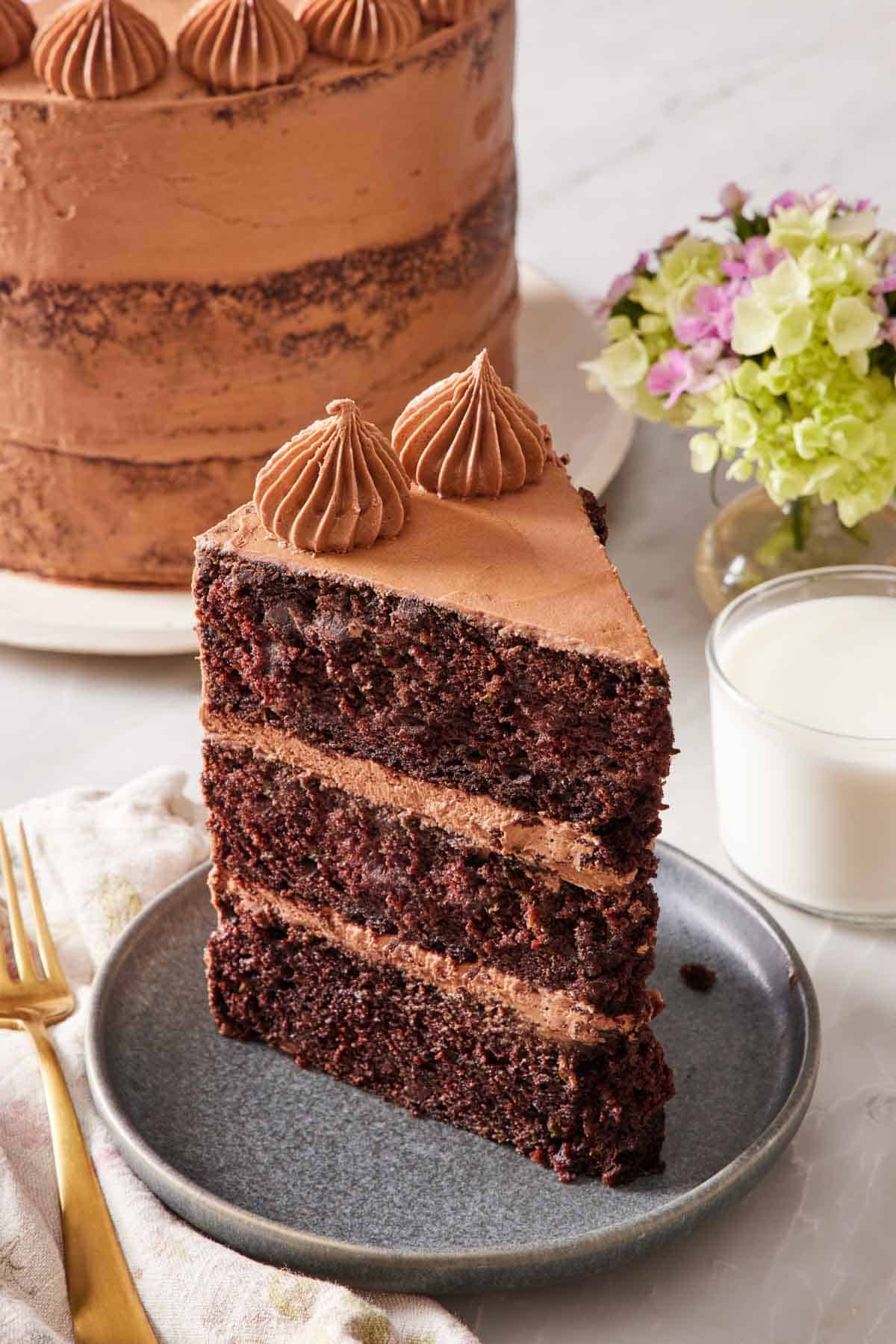 A slice of chocolate zucchini cake on a small plate with a glass of milk on the side, and the rest of the cake almost out of frame in the background.