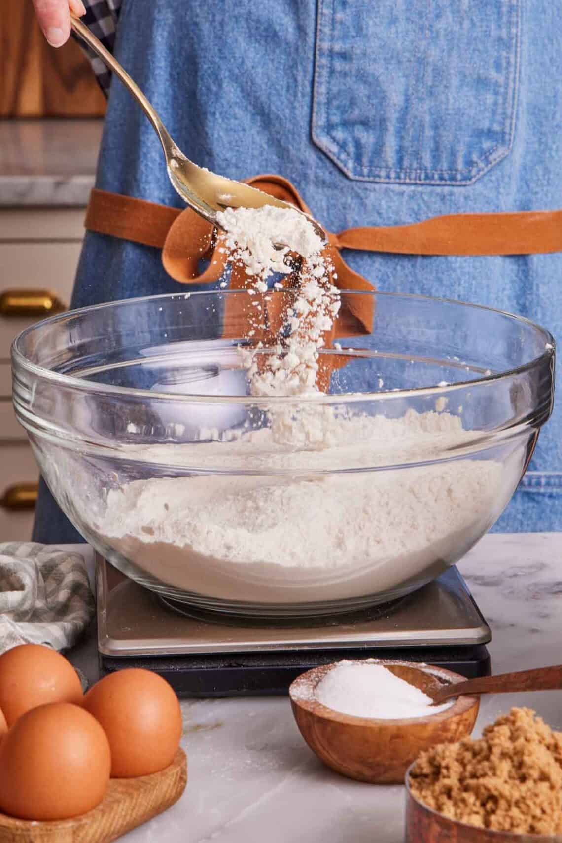 Flour spooned into a bowl containing more flour on a kitchen scale. More baking ingredients in the foreground.