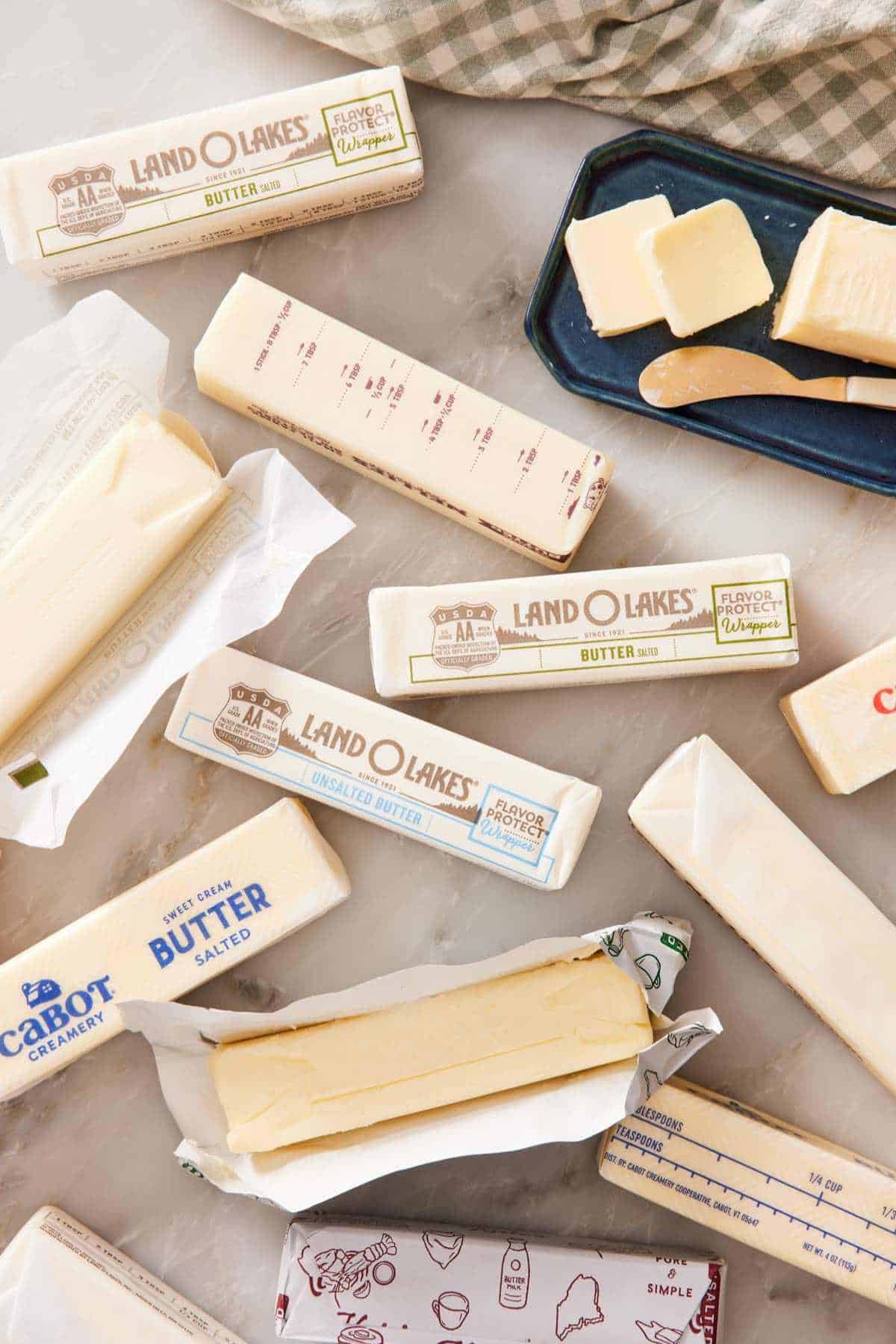 Overhead shot of different brands of sticks of butter, two unwrapped, and one stick on a butter dish with pats cut off.