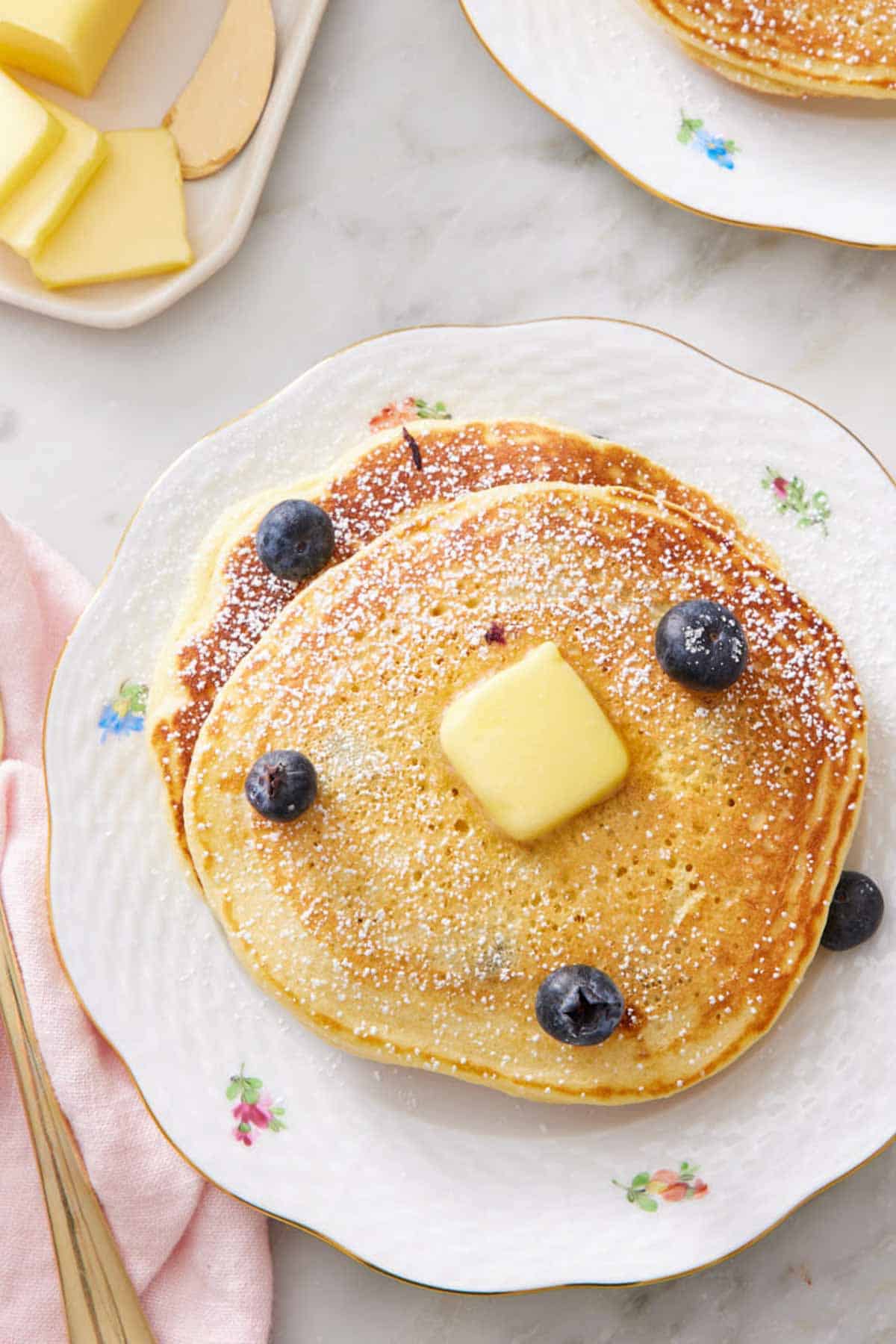 Overhead shot of blueberry pancakes on a plate with a pat of butter, powdered sugar, and blueberries on top.