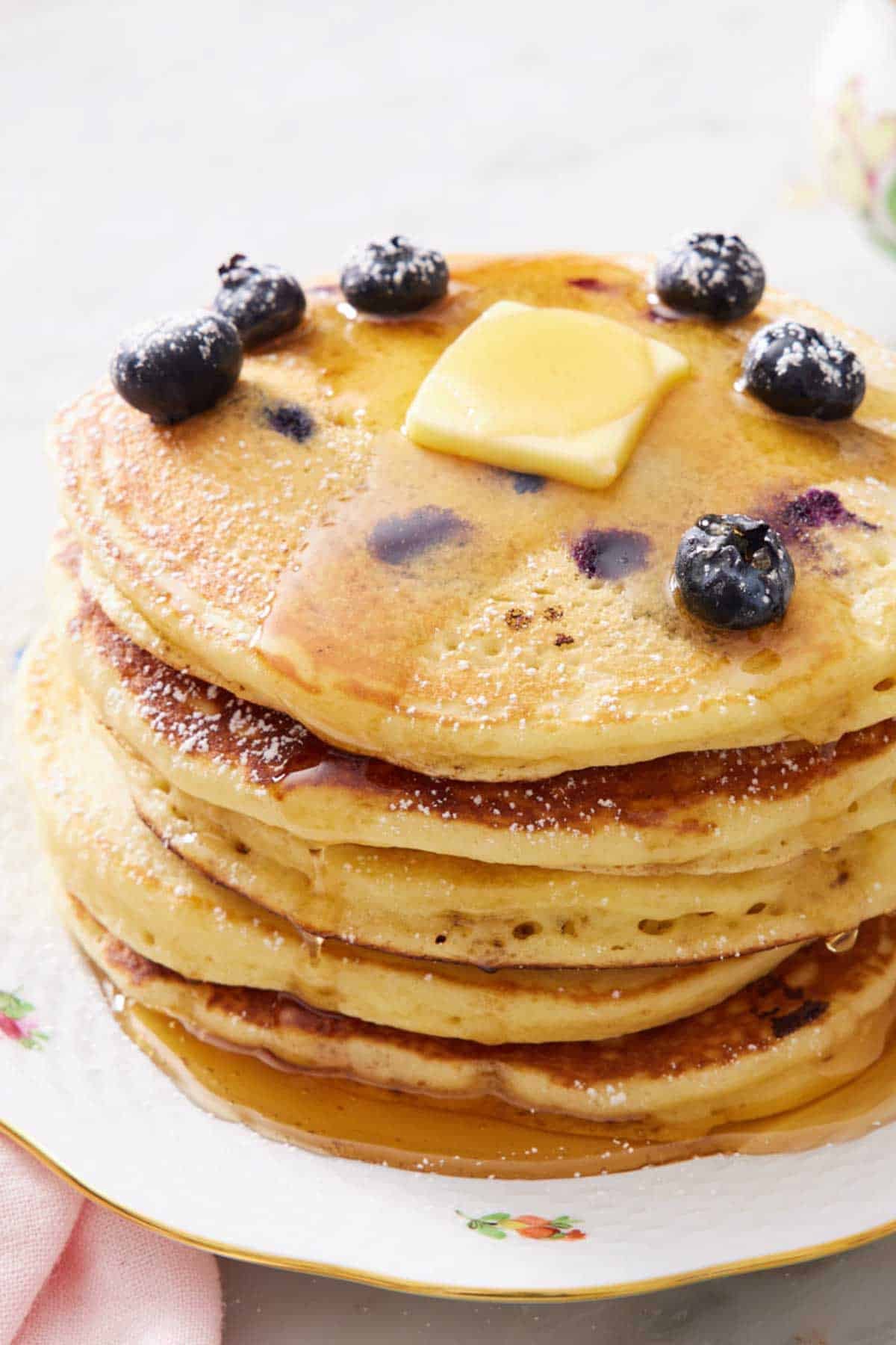 A stack of blueberry pancakes on a plat, topped with syrup, blueberries, and a pat of butter.