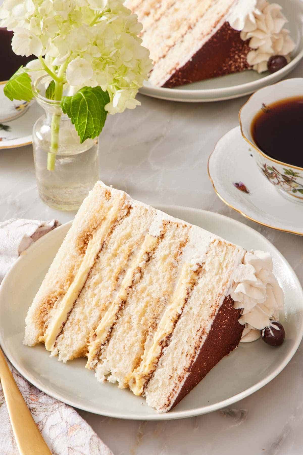 A slice of tiramisu cake on its side on a plate with a cup of coffee and another slice of cake behind it mostly out of frame.