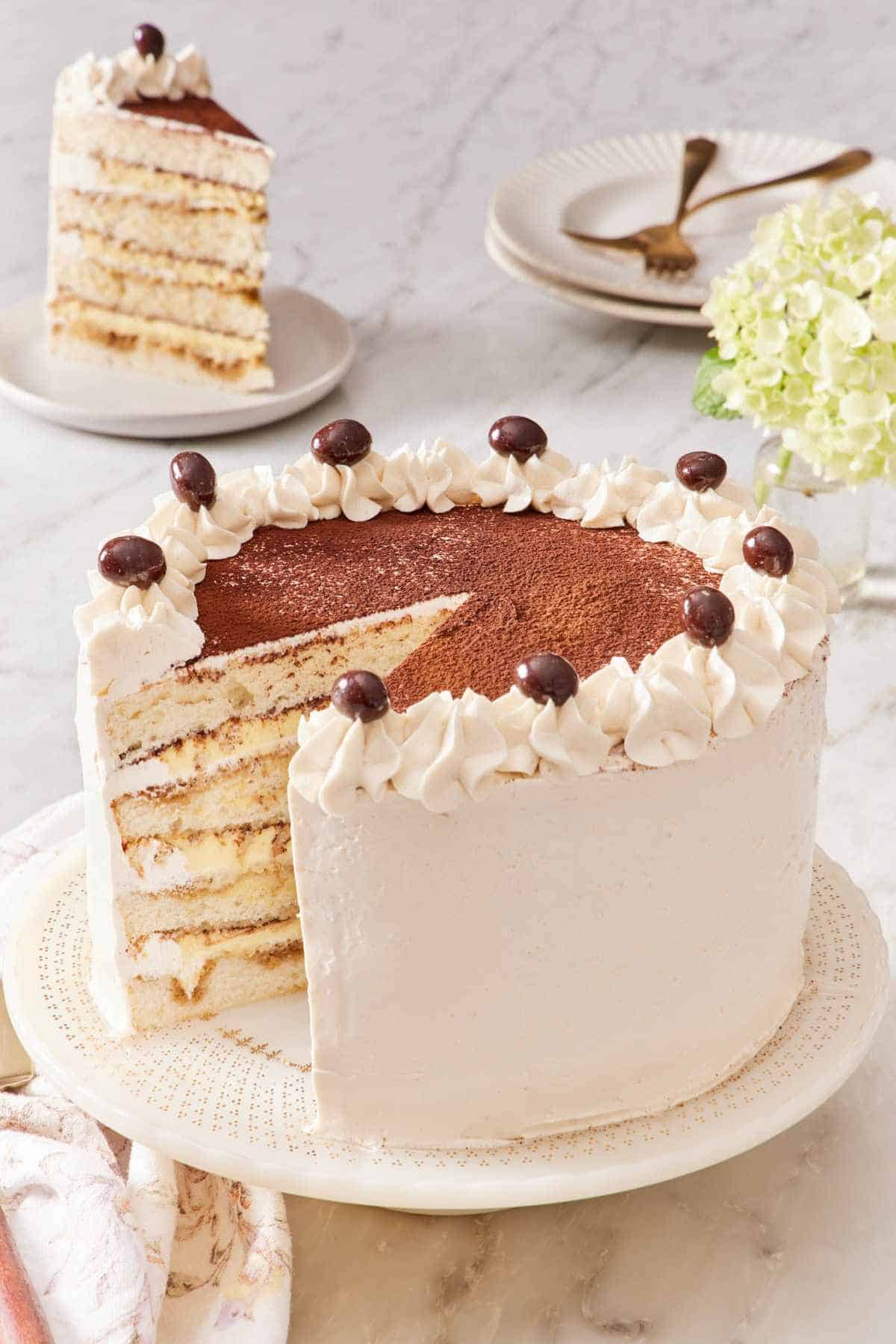Tiramisu cake on a cake stand with a slice removed and placed on a plate in the background.